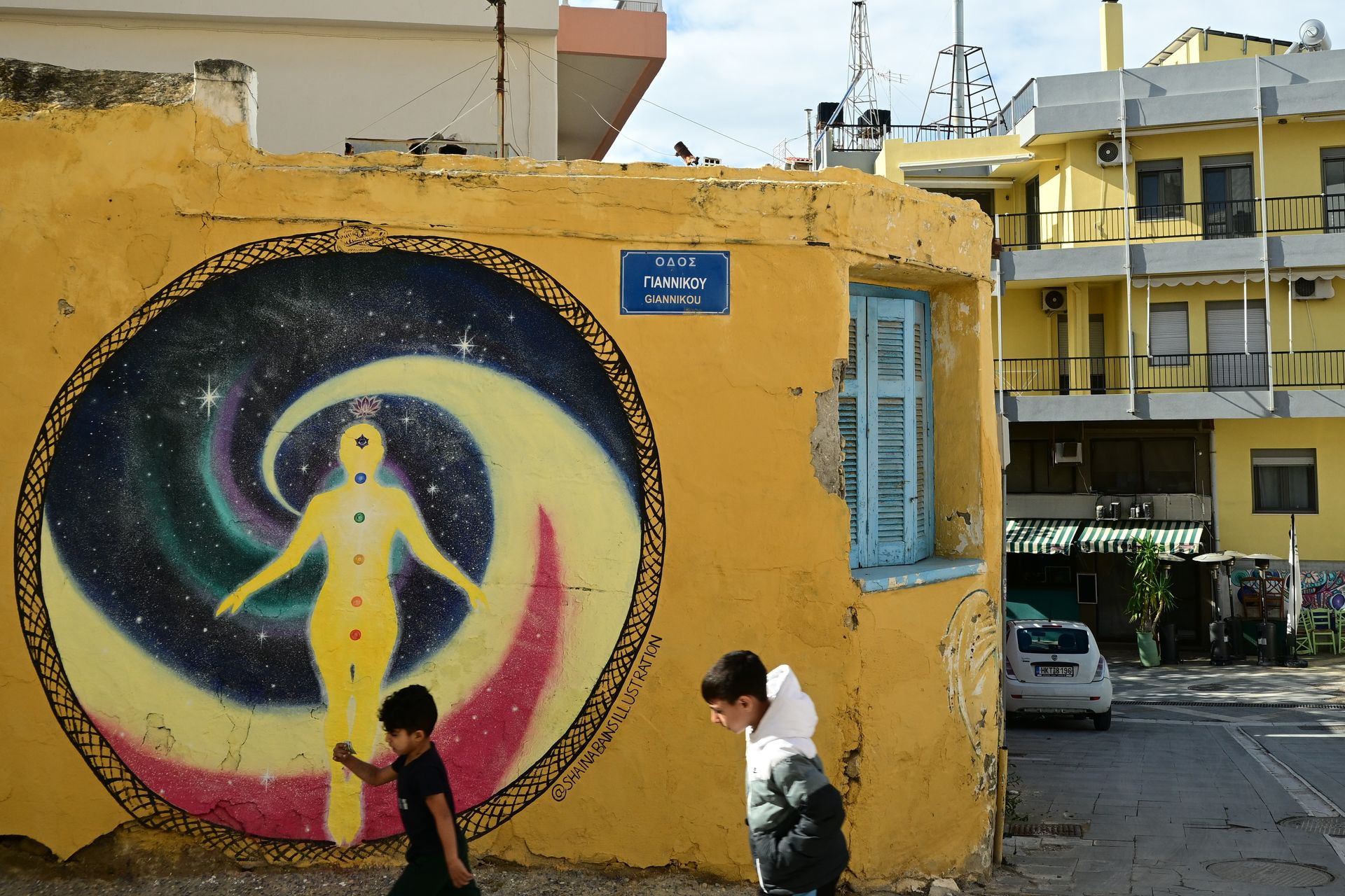 Two people walking past a circular mural on a yellow building at a street corner